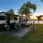 Trailer em camping brasileiro durante viagem de caravanismo, estacionado à beira de lago com estrutura de camping e área externa montada.