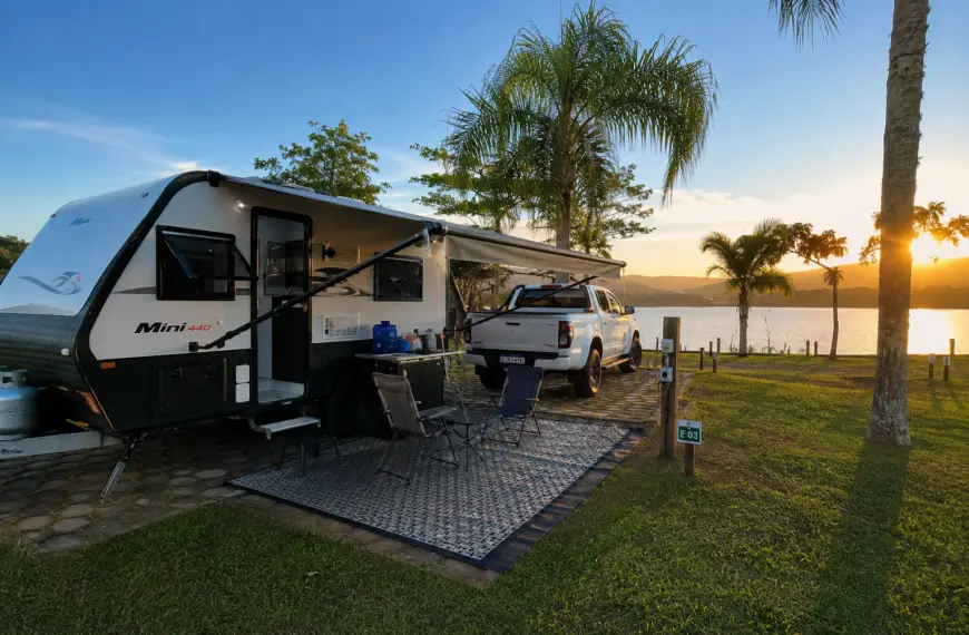 Trailer em camping brasileiro durante viagem de caravanismo, estacionado à beira de lago com estrutura de camping e área externa montada.