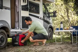 Homem ajustando gerador portátil ao lado de um motorhome estacionado em área de camping arborizada no Brasil.