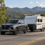 SUV Toyota cinza rebocando um trailer de viagem branco em rodovia asfaltada, cercada por vegetação verde e montanhas ao fundo.