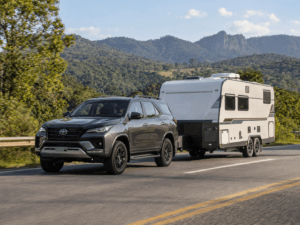 SUV Toyota cinza rebocando um trailer de viagem branco em rodovia asfaltada, cercada por vegetação verde e montanhas ao fundo.