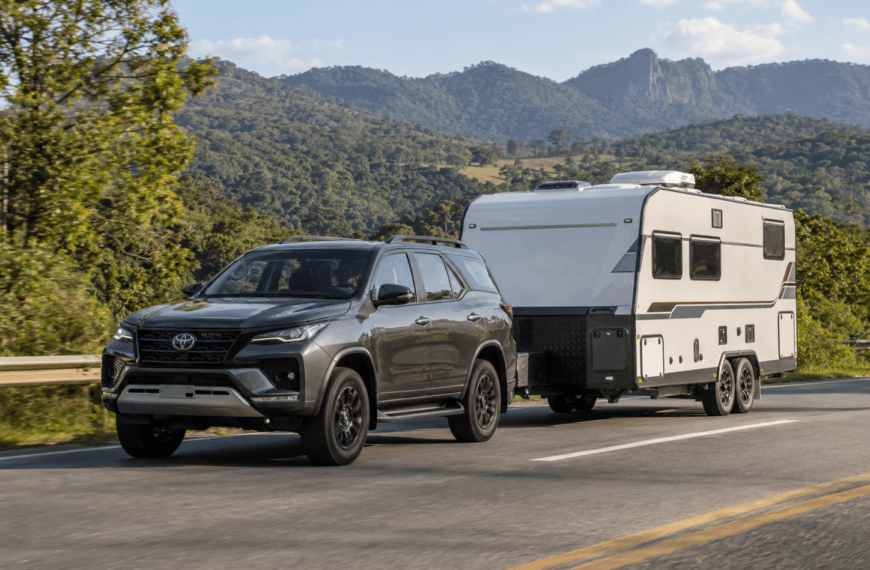 SUV Toyota cinza rebocando um trailer de viagem branco em rodovia asfaltada, cercada por vegetação verde e montanhas ao fundo.